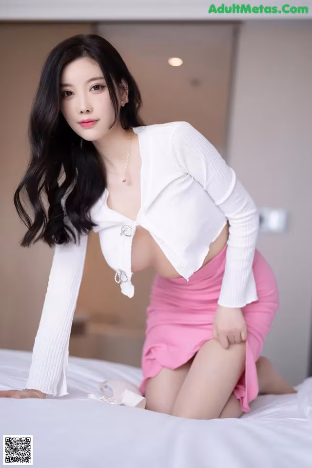 A woman in a white shirt and pink skirt sitting on a bed.