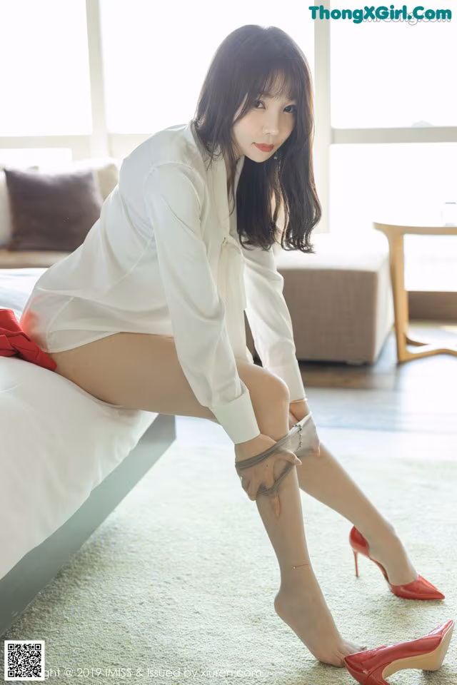 A woman in a white shirt and red shoes sitting on a bed.